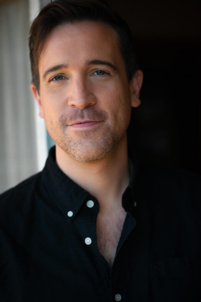 Photo of Jack Starkey-Gill, the Founder and Director of public speaking & communication consultancy JSG Voice. He is wearing a black shirt with white buttons and is smiling.